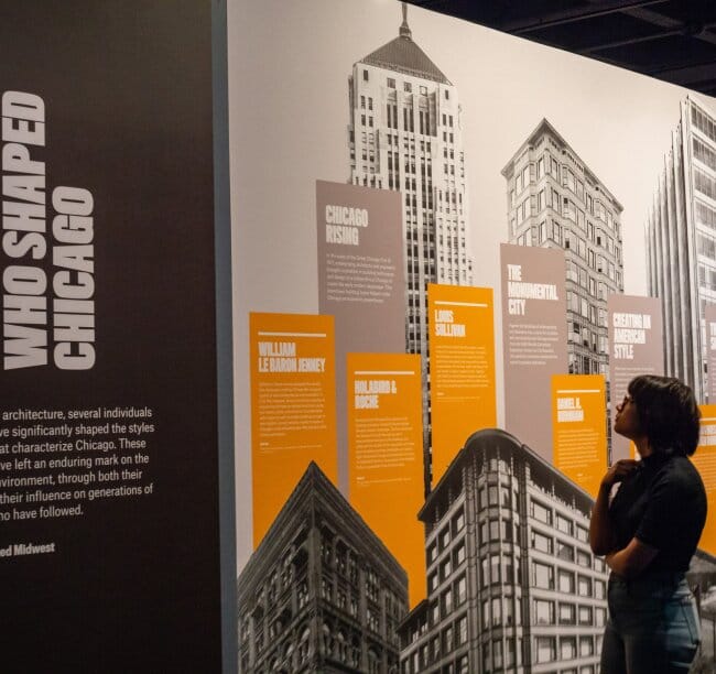 A person with a short bob looking an exhibition. The exhibition is printed on the wall with graphics of skyscrapers and order text boxes.
