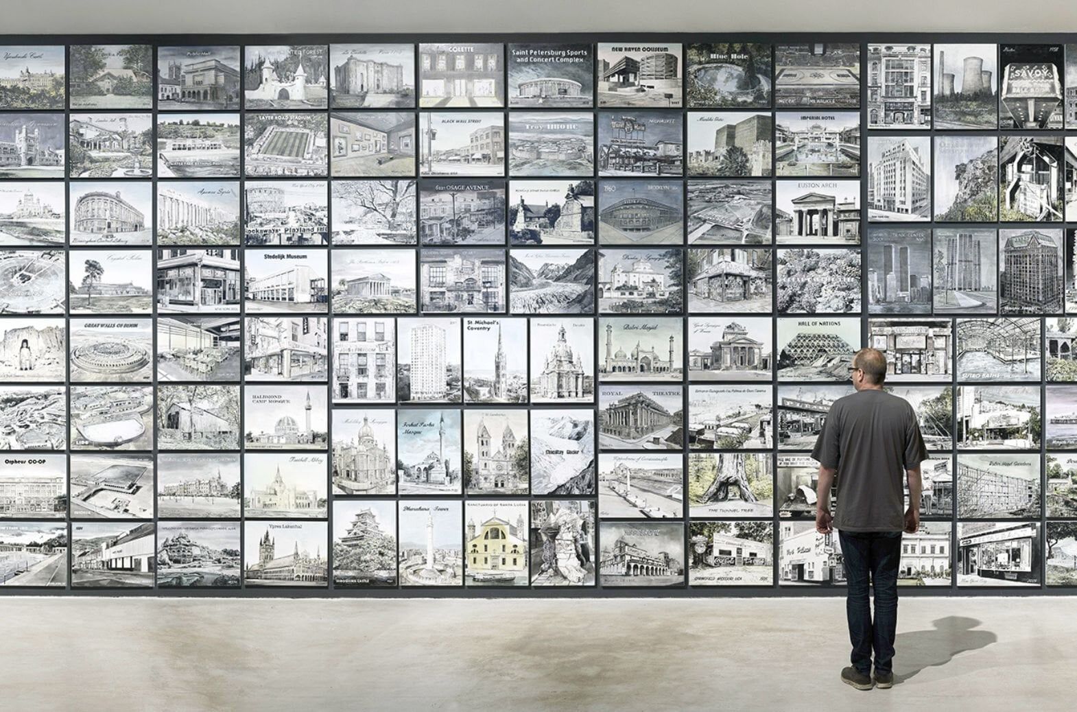 Man standing and facing away from the viewer looking at a wall of illustrations of different locations.